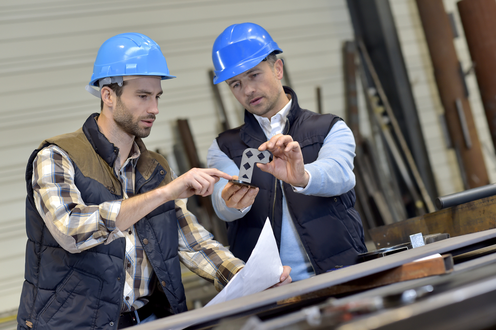Two men wearing hardhats an;yzing a small piece of machined metal.